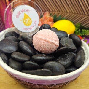 Peut inclure: Un bain moussant rose est pos&eacute; sur des pierres noires dans un panier en osier. Le panier est sur une surface en bois. Un r&eacute;cipient transparent avec le texte "Drink Drops Sips Lemonade Lush" est en arri&egrave;re-plan, avec un citron et des fleurs.