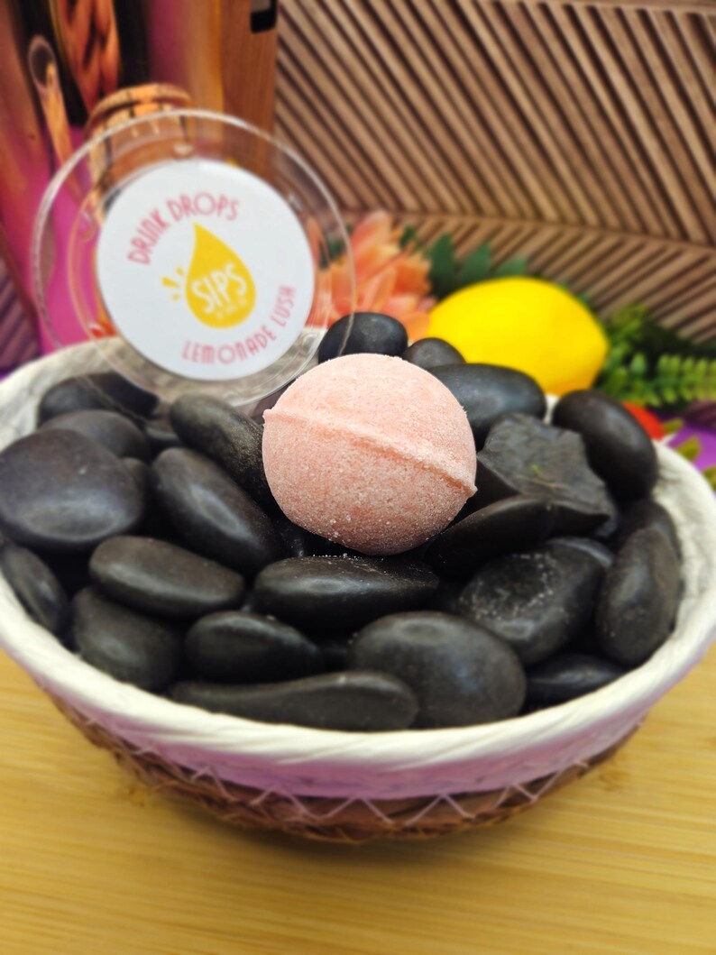 Peut inclure: Un bain moussant rose est pos&eacute; sur des pierres noires dans un panier en osier. Le panier est sur une surface en bois. Un r&eacute;cipient transparent avec le texte "Drink Drops Sips Lemonade Lush" est en arri&egrave;re-plan, avec un citron et des fleurs.