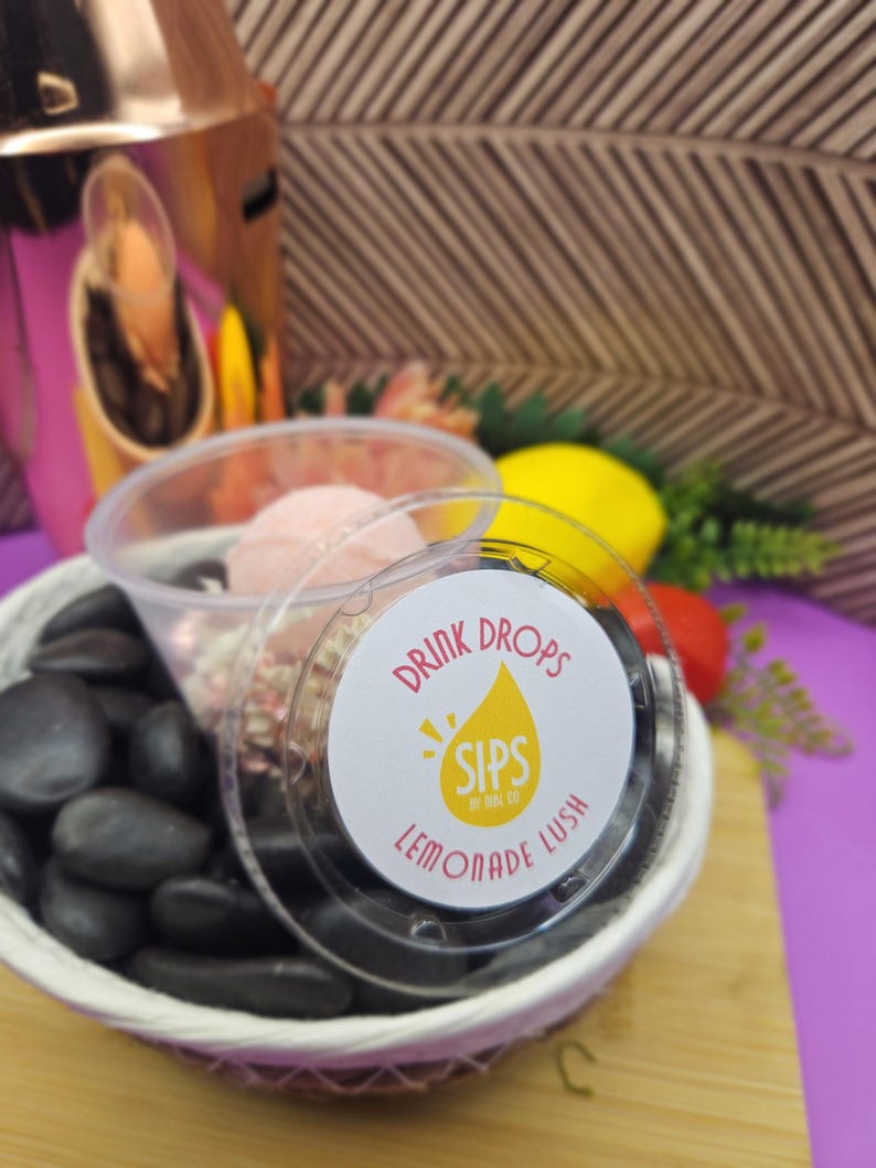 Peut inclure: Un r&eacute;cipient en plastique transparent avec une &eacute;tiquette &laquo; Drink Drops Sips by Mal Co Lemonade Lush &raquo; est pos&eacute; sur un petit panier tress&eacute; rempli de pierres noires. Une boisson rose et des vermicelles sont visibles &agrave; l'int&eacute;rieur. Un refroidisseur de boisson rose et dor&eacute; est en arri&egrave;re-plan.