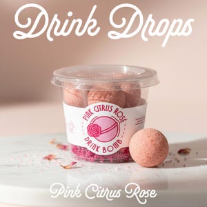 May include: A clear plastic container filled with pink, spherical drink bombs, labeled Pink Citrus Rose Drink Bomb. One individual pink, textured, spherical drink bomb sits in front of the container, surrounded by small pink floral petals and crystals.