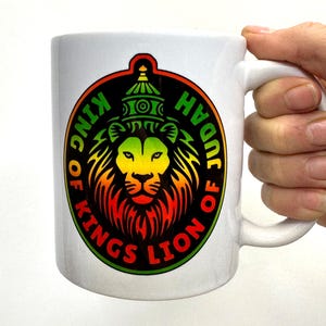 May include: White ceramic mug featuring a colorful lion design with the text "KING OF KINGS LION OF JUDAH" in a circular pattern. The lion's mane transitions from red to yellow and green. The mug is held by a hand.