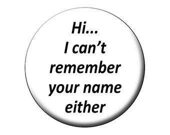 May include: A circular white pin-back button featuring the black text "Hi... I can't remember your name either" centered on the front. The button has a smooth, clean finish and a simple, bold font that is clearly legible against the white background.