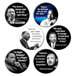 May include: A collection of six round buttons featuring portraits of Martin Luther King, Jr. Each button displays a quote attributed to him in white text against a black or blue background. The quotes address themes of justice and courage.