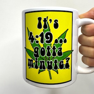 May include: White ceramic mug with a yellow rectangle featuring a green cannabis leaf and the text "It's 4:19... gotta minute?" in black. The mug has a white handle and is held by a hand.