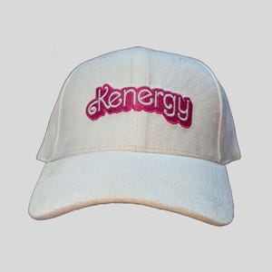 Könnte beinhalten: Weiße Baseballkappe mit einem rosa gestickten Logo, auf dem "Kenergy" steht.