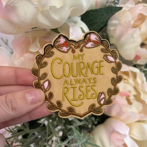 Op de afbeelding: Geborduurde patch met de tekst "MY COURAGE ALWAYS RISES" in gouddraad op een beige achtergrond. De patch heeft een decoratieve rand met roze en groene bloemaccenten. De patch is omgeven door witte en roze bloemen.