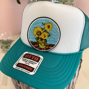 May include: A teal and white trucker hat with an embroidered patch of sunflowers in a vase. The hat has a white foam front, a teal brim, and a teal mesh back. The hat includes the text "OTTO TRUCKER".