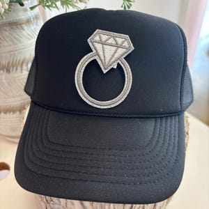 Puede incluir: Gorra de camionero negra con un parche de anillo de diamantes. El parche está bordado con un diseño de diamante y anillo gris. La gorra tiene una visera curva y una parte trasera de malla.