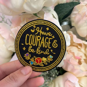 Op de afbeelding: Een ronde, zwarte geborduurde patch met gouden rand en de woorden "I have COURAGE & be kind" in gouden letters. De patch heeft kleurrijke bloemaccenten en is geplaatst tegen een achtergrond van zachte, crèmekleurige bloemen.