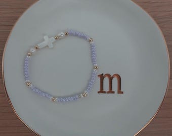 Pode incluir: Uma pulseira de contas com sementes de cor roxo claro, pequenas pérolas brancas, contas espaçadoras metálicas em tom dourado e um pingente central em forma de cruz branca, disposta em círculo sobre um prato de cerâmica branco com borda dourada.