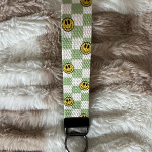 Pode incluir: Um porta-chaves de pulso xadrez verde e branco com rostos sorridentes amarelos. O porta-chaves tem um fecho de metal preto e um porta-chaves prateado. O fundo &eacute; uma superf&iacute;cie fofa, castanho claro e branca.