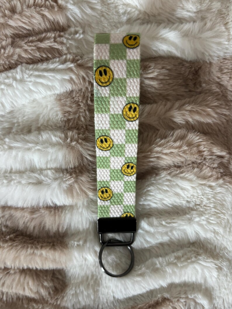 Pode incluir: Um porta-chaves de pulso xadrez verde e branco com rostos sorridentes amarelos. O porta-chaves tem um fecho de metal preto e um porta-chaves prateado. O fundo &eacute; uma superf&iacute;cie fofa, castanho claro e branca.