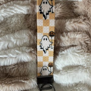 Pode incluir: Um porta-chaves de pulso com tema de Halloween com um padr&atilde;o xadrez branco e amarelo. O porta-chaves &eacute; adornado com desenhos de fantasmas e ab&oacute;boras de desenho animado. Possui um fecho de metal preto e um porta-chaves.