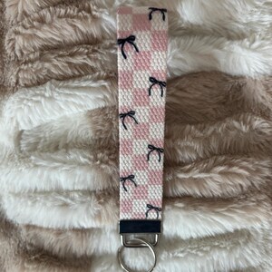 Pode incluir: Um porta-chaves de pulso xadrez rosa e branco com detalhes de la&ccedil;os pretos. O porta-chaves tem um fecho e um porta-chaves de metal prateado. O fundo &eacute; uma superf&iacute;cie fofa e texturizada, castanho claro e branco.