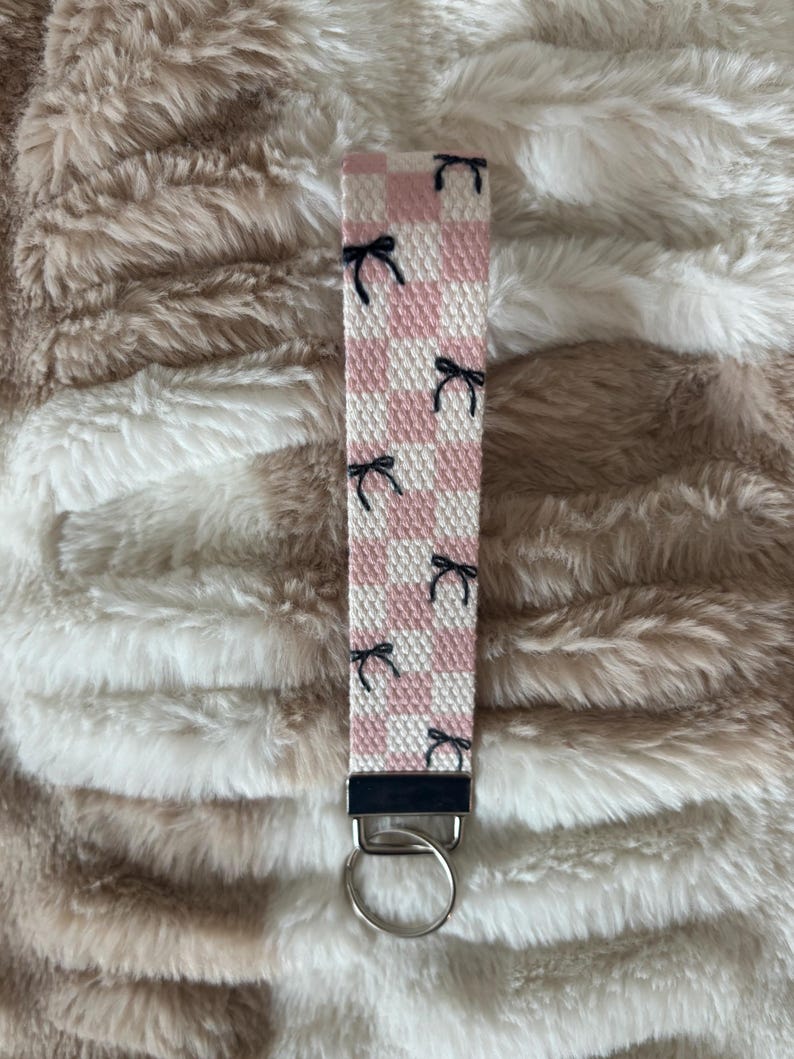 Pode incluir: Um porta-chaves de pulso xadrez rosa e branco com detalhes de la&ccedil;os pretos. O porta-chaves tem um fecho e um porta-chaves de metal prateado. O fundo &eacute; uma superf&iacute;cie fofa e texturizada, castanho claro e branco.
