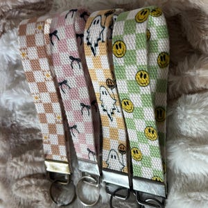 Pode incluir: Quatro chaveiros de pulso de tecido com ferragens prateadas. Os chaveiros apresentam um padr&atilde;o xadrez em v&aacute;rias cores: castanho, rosa, amarelo e verde. Os padr&otilde;es incluem la&ccedil;os, fantasmas e rostos sorridentes. Os chaveiros s&atilde;o exibidos em uma superf&iacute;cie fofa e clara.