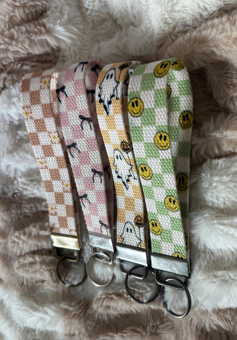 Pode incluir: Quatro chaveiros de pulso de tecido com ferragens prateadas. Os chaveiros apresentam um padr&atilde;o xadrez em v&aacute;rias cores: castanho, rosa, amarelo e verde. Os padr&otilde;es incluem la&ccedil;os, fantasmas e rostos sorridentes. Os chaveiros s&atilde;o exibidos em uma superf&iacute;cie fofa e clara.