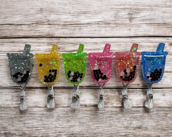Pode incluir: Seis porta-crachás coloridos com glitter em forma de chá de bolhas. Cada copo tem uma cor diferente: cinza, amarelo, verde, rosa, vermelho e azul. Os copos têm contas pretas e um canudo. Os porta-crachás são presos a um clipe prateado.
