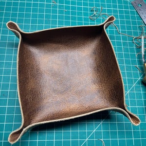 May include: A square, brown leather tray with rounded corners and raised edges. The tray is made of textured, distressed leather. Tools, including a ruler and a knife, are visible in the background.