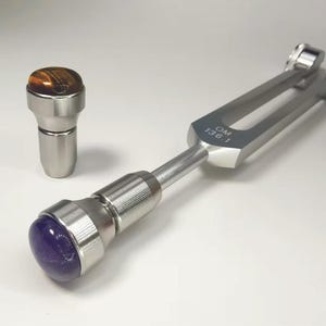 Puede incluir: Un diapasón plateado con el texto "OM 136.1" y una varita a juego con una parte superior de cristal. La varita tiene un cristal de amatista púrpura. Una segunda varita tiene un cristal de ojo de tigre. El diapasón es una herramienta para la curación por sonido.