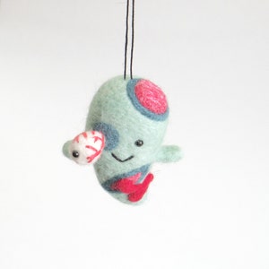 May include: A green felt ornament with red and blue details, a white eye, and a smiling face. The ornament is hanging from a black string.