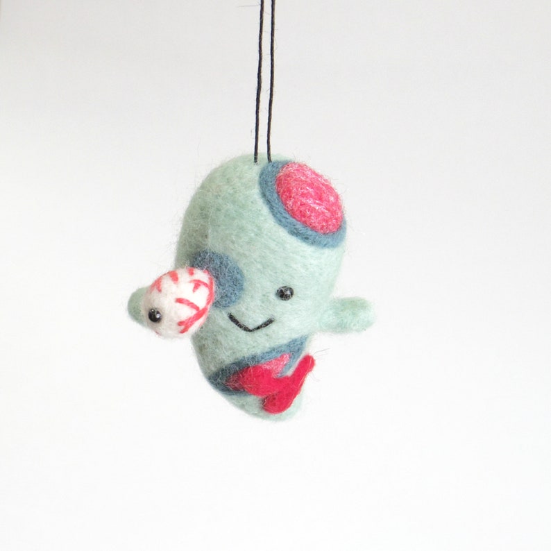 May include: A green felt ornament with red and blue details, a white eye, and a smiling face. The ornament is hanging from a black string.