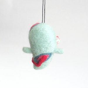 May include: A teal blue felt ornament with red and white accents. The ornament is shaped like a bird with a small beak and a tail.