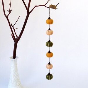 May include: A string of felt pumpkins in shades of green, orange, and cream hangs from a branch. The pumpkins are strung together with twine and have black stems. The tag reads "fuzz & frills".