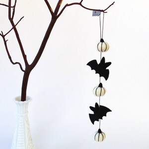 May include: A white felt garland with black bats and white pumpkins hanging from a branch. The garland is perfect for Halloween decor.