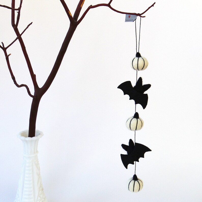 May include: A white felt garland with black bats and white pumpkins hanging from a branch. The garland is perfect for Halloween decor.
