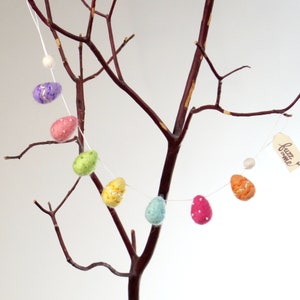 以下が含まれることがあります： 紫、ピンク、緑、黄、青、オレンジなど、さまざまな色のフェルト製のイースターエッグのガーランドが、裸の枝からぶら下がっています。卵は白いドットで飾られており、いくつかはキラキラしています。ガーランドには「fuzz & me」というラベルが付いています。