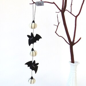 May include: A felt Halloween garland with three white pumpkins and two black bats hanging from a string. The garland is hanging from a branch in a vase.