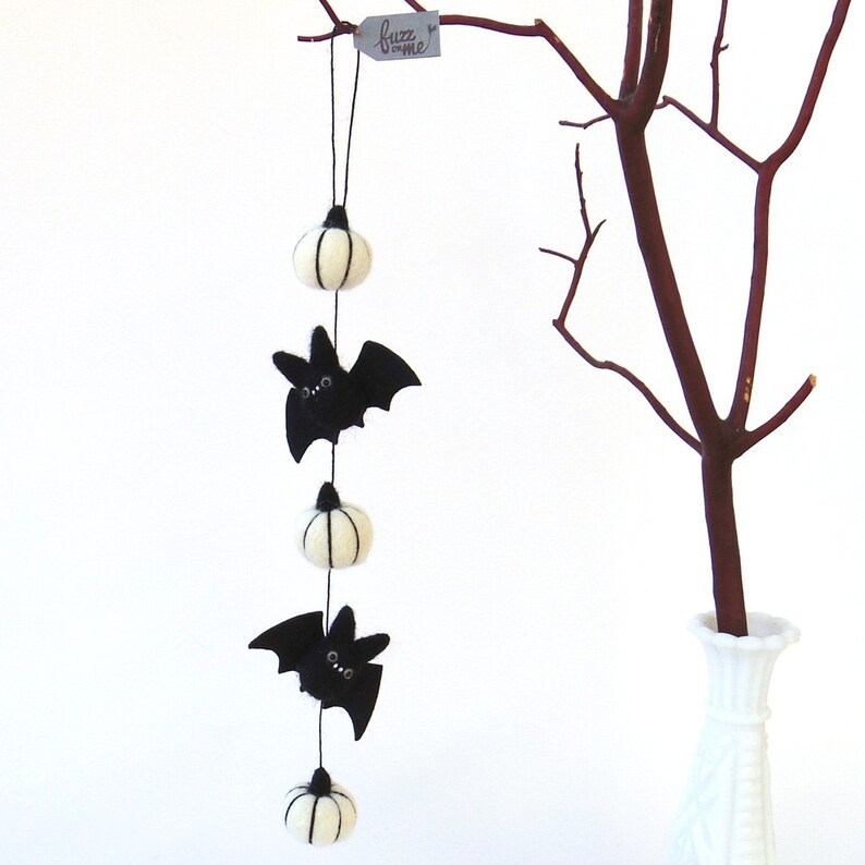 May include: A felt Halloween garland with three white pumpkins and two black bats hanging from a string. The garland is hanging from a branch in a vase.