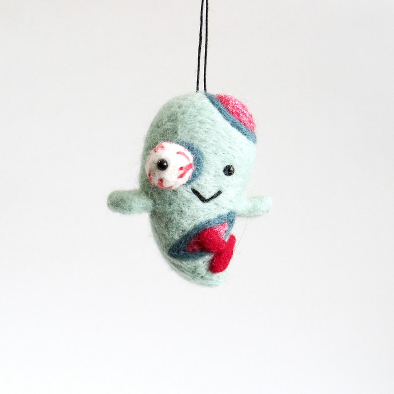 May include: A green felt ornament shaped like a monster with a single eye, a red mouth, and a red and blue design. The ornament is hanging from a black string.