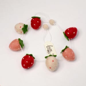 以下が含まれることがあります： ピンク、赤、白のイチゴのフェルトガーランド。ガーランドは白い紐に通されており、小さなタグには「buzz chime」と書かれています。