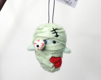 May include: A handmade felt mummy ornament with a light green wrap, a red heart, and a detached eyeball. The mummy has black stitched details and a tag that says "fuzz on me".