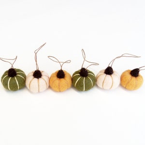May include: A row of six felt pumpkins in various colors: olive green, cream, and shades of orange. Each pumpkin has a dark brown stem and a brown string for hanging. The pumpkins are arranged on a white background.