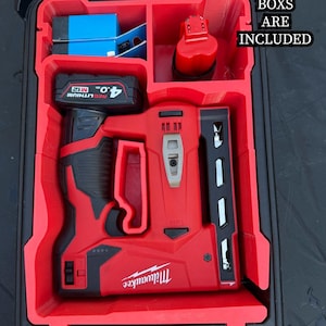 May include: A red Milwaukee M12 cordless nailer and battery pack are stored in a red Packout organiser. The organiser has compartments for tools and accessories. Text on the image reads: "NO TOOLS OR PACKOUT BOXES ARE INCLUDED."