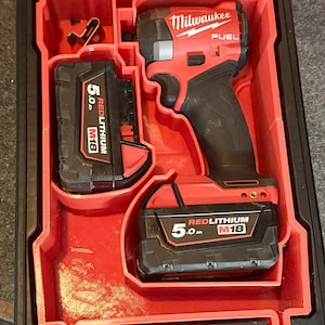 Peut inclure: Un kit de visseuse à chocs Milwaukee FUEL rouge et noir. Le kit comprend la visseuse à chocs, deux batteries REDLITHIUM M18 de 5,0 Ah et un coffret de rangement. Le logo Milwaukee est visible.