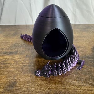 May include: A dark purple, egg-shaped decorative object with a large, teardrop-shaped opening. A matching purple, spiky-tailed creature is positioned below the egg. The object is on a wooden surface.