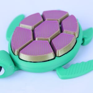 Op de afbeelding: Een groen plastic schildpad speelgoed met een roze en gouden schild. Het schild is gemaakt van meerdere zeshoekige stukken.