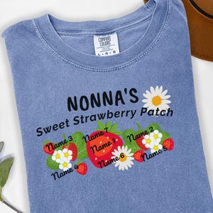 May include: Blue jean colored t-shirt with the text "NONNA'S Sweet Strawberry Patch". The design includes a large red strawberry, daisies, and smaller strawberries with names. The shirt is a Comfort Colors brand.