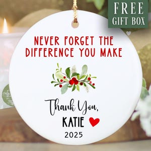May include: A white ceramic ornament with the text "NEVER FORGET THE DIFFERENCE YOU MAKE" in red. Below is "Thank You, KATIE" and a red heart. A floral design with red berries and green leaves is in the centre. The year 2025 is at the bottom.