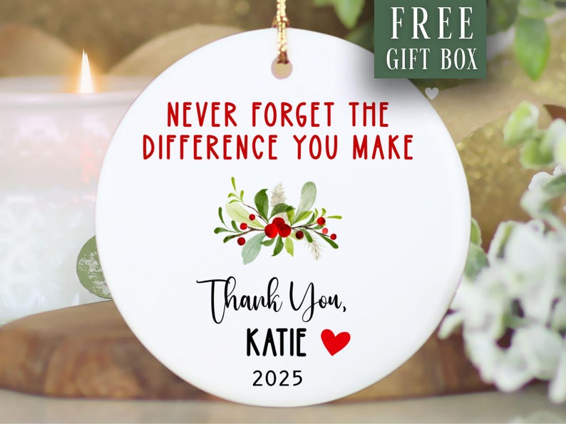 May include: A white ceramic ornament with the text "NEVER FORGET THE DIFFERENCE YOU MAKE" in red. Below is "Thank You, KATIE" and a red heart. A floral design with red berries and green leaves is in the centre. The year 2025 is at the bottom.