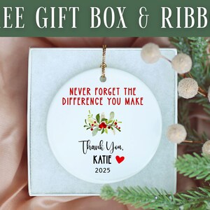 May include: A white ceramic ornament with the text "NEVER FORGET THE DIFFERENCE YOU MAKE" in red. Below, it reads "Thank You, KATIE" with a red heart and the year "2025". The ornament is in a gift box with a sprig of greenery.