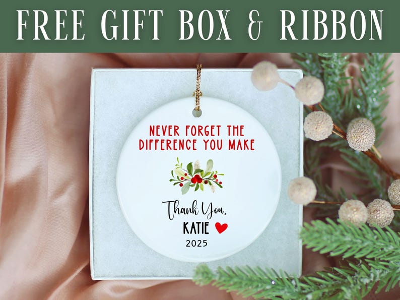 May include: A white ceramic ornament with the text "NEVER FORGET THE DIFFERENCE YOU MAKE" in red. Below, it reads "Thank You, KATIE" with a red heart and the year "2025". The ornament is in a gift box with a sprig of greenery.