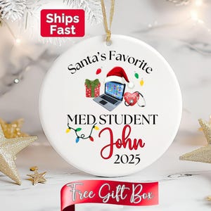 May include: A white ceramic Christmas ornament with a gold string for hanging. The ornament features the text "Santa's Favorite MED STUDENT John 2025" with illustrations of a laptop, Santa hat, gift, and stethoscope. A red banner at the bottom reads "Free Gift Box".