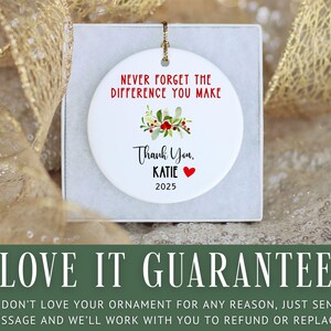 May include: White ceramic ornament with a gold string, inscribed with "NEVER FORGET THE DIFFERENCE YOU MAKE." It has a floral design, the words "Thank You, KATIE," a red heart, and the year 2025. The ornament is presented in a white box.