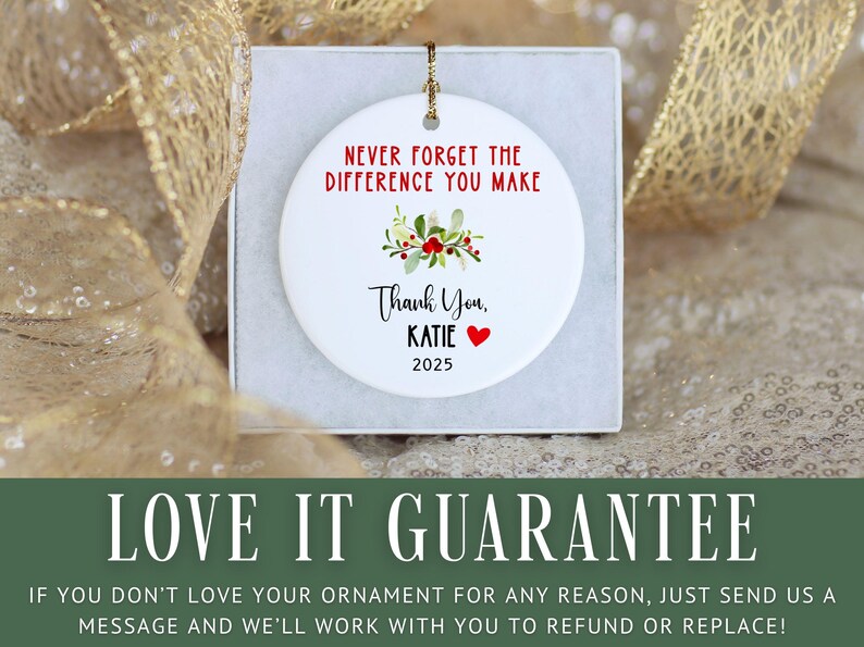May include: White ceramic ornament with a gold string, inscribed with "NEVER FORGET THE DIFFERENCE YOU MAKE." It has a floral design, the words "Thank You, KATIE," a red heart, and the year 2025. The ornament is presented in a white box.