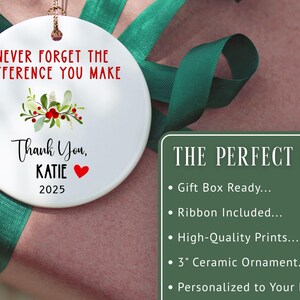 May include: A white ceramic ornament with the text "NEVER FORGET THE DIFFERENCE YOU MAKE" and "Thank You, KATIE 2025". The ornament has a floral design and a red heart. The ornament is approximately 7.6 cm in diameter.